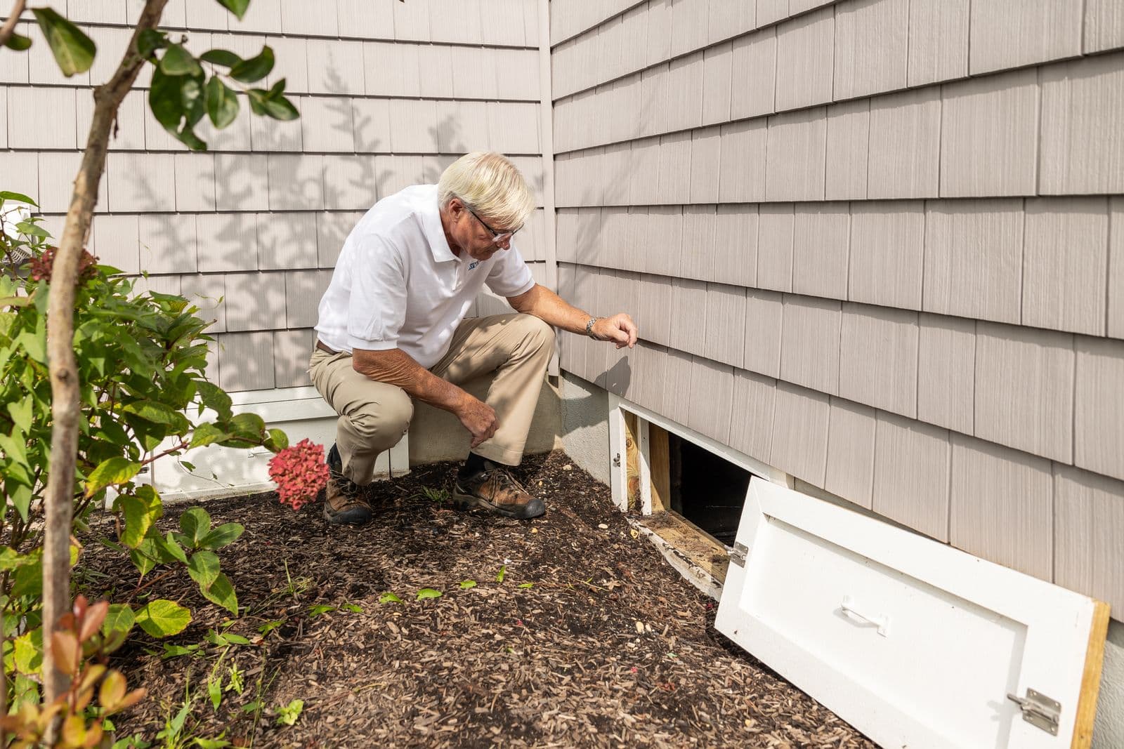 Inspector crouching at an open crawl space access door on the side of a house